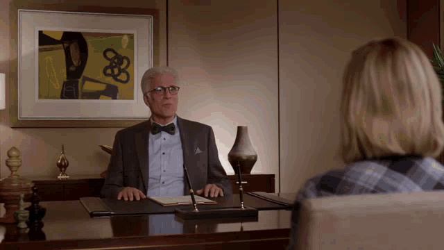 a man in a bow tie sits at a desk in front of a framed picture